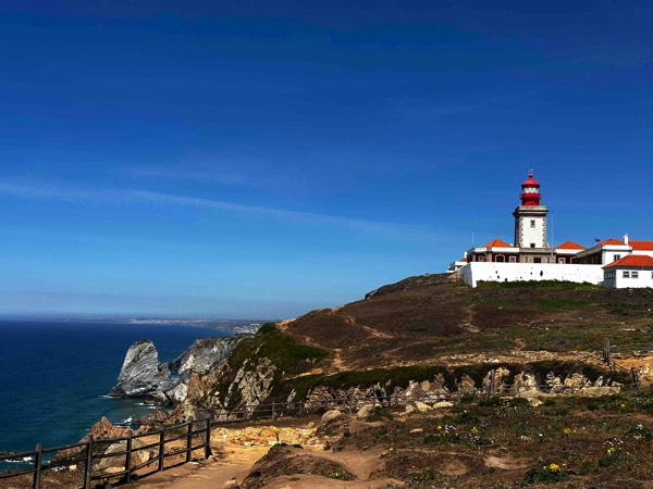 Photo of a red and white lighthouse sitting on a promontory overlooking the blue Atlantic Ocean against blue skies