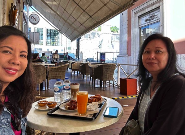 Two women having lunch with a round table between them filled with plates of patries and beverages at an outdoor cafe